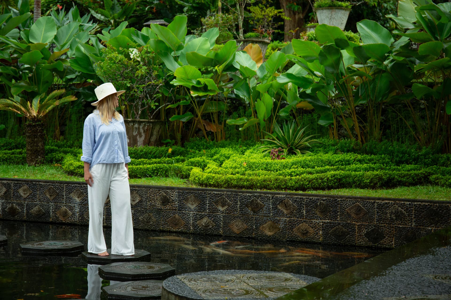A gardener in a sun hat standing on stepping stones above a koi pond, surrounded by broad tropical foliage and clipped hedging